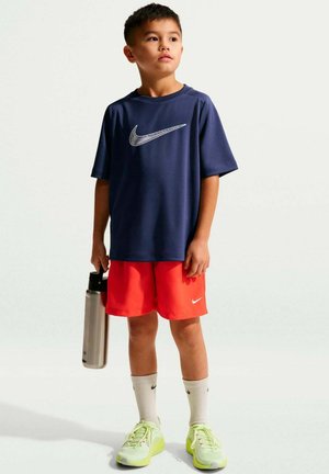 Ragazzo giovane che indossa una maglietta Nike blu navy, pantaloncini rossi, calzini bianchi e scarpe da ginnastica gialle, che tiene una bottiglia d'acqua argento, in piedi su uno sfondo bianco.