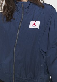 Granatowa kurtka przeciwwiatrowa z wysokim kołnierzem, zapinana na zamek, z elastycznymi mankietami. Posiada białą naszywkę z czerwonym logo na piersi.