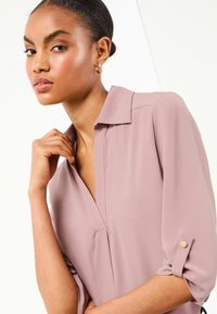 Blusa rosa chiaro con scollo a V e maniche arrotolate, caratterizzata da dettagli con bottoni in tonalità dorata. Il tessuto appare liscio con una leggera lucentezza.