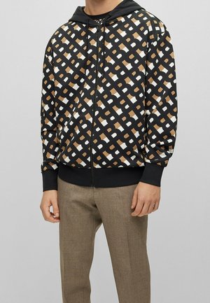 Mies, joka pitää mustaa vetoketjullista hupparia, jossa on beige ja valkoinen geometrinen ketjumalli, sekä beiget housut, seisoo vaalean harmaan taustan edessä.