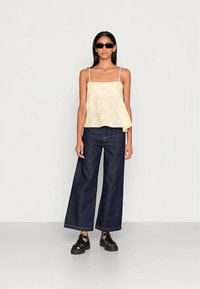 Top amarelo claro de alças finas com uma silhueta evasê e bolinhas brancas, combinado com jeans escuro de perna larga e sandálias pretas.