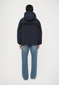 Casaco puffer azul-marinho com capuz, apresentando uma faixa preta nas costas e um tecido texturizado. Combinado com jeans de perna larga azul-clara.