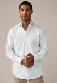 Hvid skjorte med knapper lavet af glat stof, med en klassisk krave, lange ærmer og en buet kant. Hænderne lagt sammen foran.