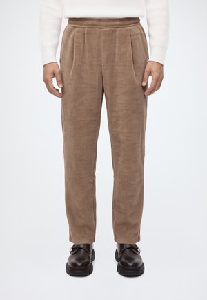 Personne portant un pantalon en velours côtelé beige, des chaussures en cuir noires et un pull blanc côtelé, debout sur un fond blanc uni.