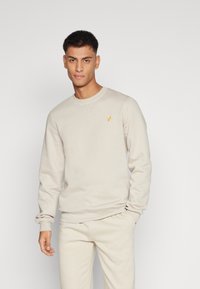 Beige crewneck sweatshirts med lange ærmer, med et lille gult logo på brystet, lavet af blødt stof med en glat tekstur.
