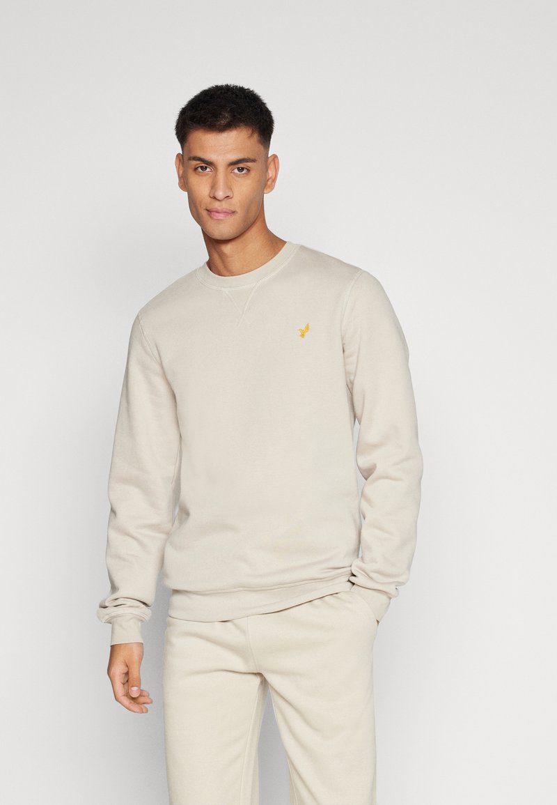 Sweatshirt beige à col rond avec manches longues, doté d'un petit logo jaune sur la poitrine, fabriqué en tissu doux avec une texture lisse.