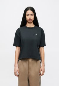 Schwarzes, kurz geschnittenes T-Shirt aus weichem Stoff mit kurzen Ärmeln, versehen mit einem kleinen weißen "obey"-Logo auf der Brust; kombiniert mit beigen Hosen.