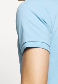 Person som bär en ljusblå kortärmad skjorta, som visar överarmen och axelområdet mot en enfärgad bakgrund.