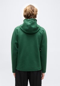  Άτομο με κοντά ξανθά μαλλιά στέκεται με την πλάτη, φορώντας μια σκούρα πράσινη κουκούλα και μαύρο παντελόνι σε απλό ανοιχτό φόντο.