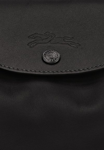 Schwarze Lederhandtasche mit Klappe, die ein geprägtes Greyhound-Logo und einen runden Metallknopf mit gestempeltem Markennamen und dem Jahr 1948 aufweist.