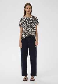 Noor naine seisab, kandes beeži ja musta mustriga lühikese varrukaga topi, tumedaid laia säärega teksaseid ja musti sandaale, ühevärvilise hele tausta ees.