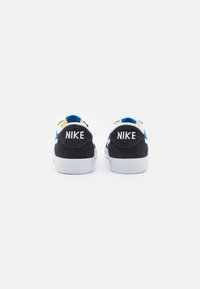 Melnas zamšādas un sieta sporta apavus ar baltu zoli, zilām un dzeltenām akcentiem, un "NIKE" logotipu uz papēža. Priekšā plakana auklu aizdare.