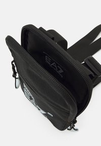 Borsa a tracolla nera in nylon con chiusura a zip, dotata di un patch con logo, tracolla regolabile e superficie testurizzata. Visibili più scomparti.