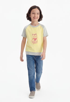 Sretno dijete hoda, nosi žutu i sivu kratku majicu s ružičastim likom čudovišta i plave traperice, na bijeloj pozadini.