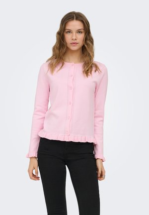 Jeune femme portant un cardigan rose clair boutonné avec des bords volantés et un pantalon noir, debout devant un fond clair uni.