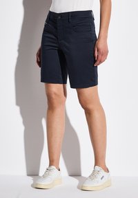 Pantalones cortos azul marino de corte recto con dos bolsillos laterales. Presentan un cierre de botón y un dobladillo cosido para mayor durabilidad. Se llevan con zapatillas blancas.