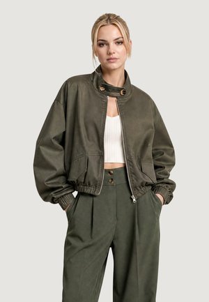 Femme portant une veste courte vert olive avec un col boutonné et un pantalon taille haute assorti, debout les mains dans les poches.