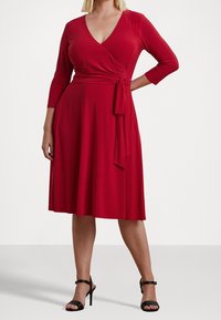 Vestido envolvente rojo de longitud a la rodilla, confeccionado en una tela suave, con escote en V y mangas tres cuartos, que presenta un cinturón anudado en la cintura. Sandalias negras de tacón alto.