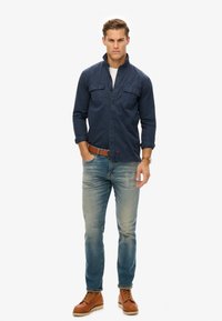 Camisa de botones azul marino con dos bolsillos en el pecho, jeans celestes y botas marrones. El modelo está de pie con una expresión neutral.