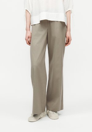 Personne portant un pantalon ample beige, un haut texturé blanc à manches mi-longues, et des baskets claires à lacets, debout sur un fond blanc.