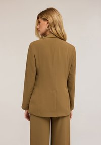 Hnědý přizpůsobený blejzr se strukturovanými rameny, dlouhými rukávy a hladkou texturou, spárovaný s odpovídajícími kalhotami.