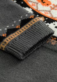 Pull gris tricoté avec un poignet texturé présentant un motif complexe en noir, orange et blanc. Détails côtelés accentuant la manche.