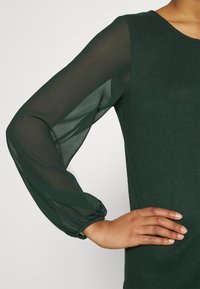 Grünes strukturiertes Oberteil mit transparenten, gepufften Ärmeln. Die Ärmel sind leicht durchsichtig und haben einen gerafften Ärmelbund. Strukturierter Oberkörper.