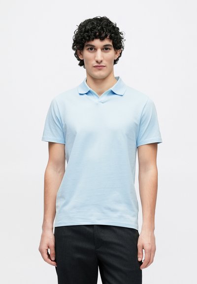 Gaiši zilas krāsas polo krekls ar īsām piedurknēm un apkakli, izgatavots no gludas auduma. Valkāts ar tumšām, teksturētām biksēm. Vienkāršs dizains, bez rakstiem.