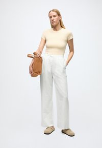 Beige figursydd t-shirt, vita vida byxor och beigea slip-on skor. Håller en liten brun mockaväska med två handtag. Minimalistisk design.