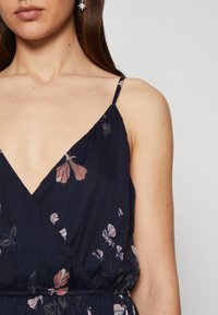 Blommig byxdress med marin bakgrund, prydd med delikata rosa och vita blommor, tunna justerbara axelband och V-ringning. Slät tygkvalitet.