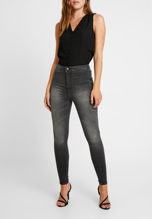 Vrouw die een mouwloze zwarte blouse met knopen, vervaagde zwarte skinny jeans en zwarte high-heeled sandalen met bandjes draagt, staand tegen een witte achtergrond.