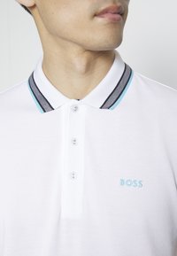 Homme portant un polo blanc à trois boutons avec col rayé, logo "BOSS" bleu brodé sur la poitrine.