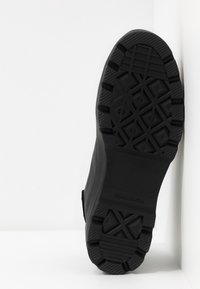 Semelle en caoutchouc noir d'une botte debout verticalement sur un fond blanc avec des motifs de bande de roulement profonds et le nom de la marque Converse.