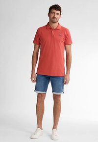 Rotes Baumwoll-Poloshirt mit Kragen und Logo, kombiniert mit Jeansshorts. Das Modell trägt weiße Sneaker und zeigt einen neutralen Gesichtsausdruck.