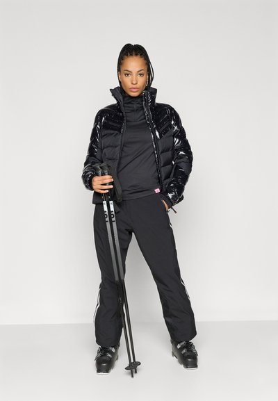 Casaco puffer preto com acabamento brilhante, gola alta, design em camadas, combinado com calças de ski pretas, segurando bastões de ski pretos, calçado com botas de neve.