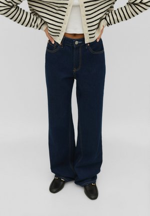 Persoon die hoge, wijd uitlopende donkerblauwe spijkerbroek draagt, zwarte schoenen, een wit cropped top en een crème gebreid vest met zwarte strepen, staand met de handen in de zij.