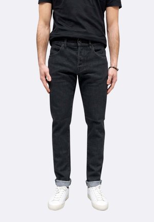 Uomo con jeans neri slim fit risvoltati alle caviglie, scarpe da ginnastica bianche, t-shirt nera e un braccialetto al polso destro.