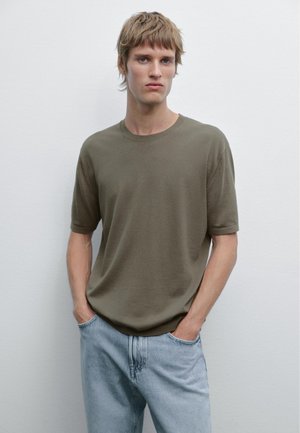 Un giovane modello maschio sta in piedi con le mani in tasca, indossando una camicia verde e jeans azzurri chiari su uno sfondo bianco semplice.