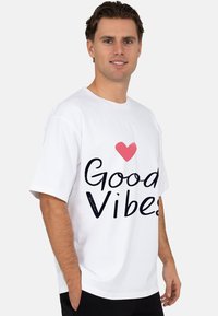 Witte katoenen t-shirt met een zwarte "Good Vibes" opdruk en een roze hartaccent. Korte mouwen en een losse pasvorm.