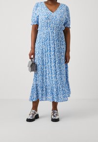 Kvinna i en blå och vit blommig midiklänning, som håller en grå handväska och har på sig glänsande silverfärgade skor med spännen.