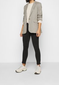 Beige und schwarze Hahnentrittmuster-Jacke, getragen über einem cremefarbenen Kabelstrick-Pullover, kombiniert mit schwarzen Skinny Jeans und hellen Sneakers.