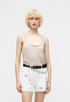 Bleg person med krøllet hår iført en lysebeige tanktop, hvide shorts med blomsterbroderi, brun fletbælte og sølvkædebælte.