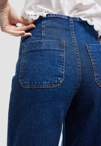 Höga blå denimjeans med en slät textur, som har två bakfickor och kontrasterande orange sömmar.