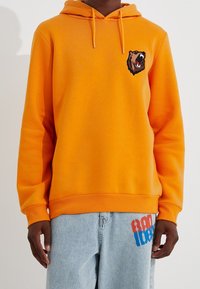 Sudadera naranja hecha de tejido suave, con un gráfico de un oso en el pecho. El diseño incluye una capucha con cordón y puños acanalados.