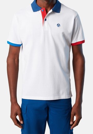 Man wearing a white polo shirt with a blue collar, red placket, blue and red sleeve cuffs, and blue shorts with cargo pockets.
