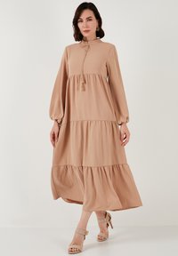 Beige dlouhé šaty s vysokým límcem, dlouhými balónovými rukávy a vrstvenou sukni. Má stáhnutí na šňůrku se třásněmi. Hladká textura.