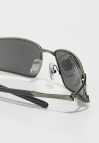 Lunettes de soleil avec des verres teintés gris, une monture métallique verte, des repose-nez en caoutchouc souple et des branches bleu foncé avec le logo "ARNETTE".