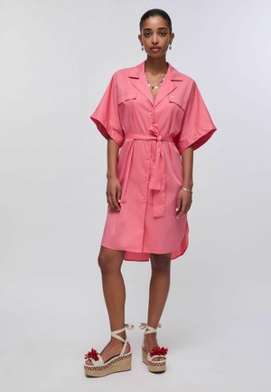 Femme portant une robe-chemise rose ceinturée mi-longue à manches courtes, associée à des sandales beiges à plateforme décorées d'accents rouges.