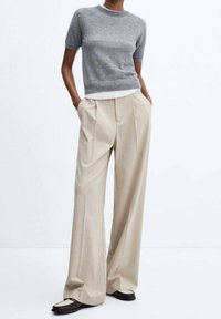 Pull en maille gris à manches courtes associé à un pantalon ample plissé beige et des chaussures marron foncé à enfiler avec des accents blancs.