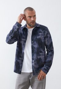 Camisa de corduroy en color azul marino con efecto tie-dye, con costillas verticales, cierre de botones en la parte delantera y cuello puntiagudo, superpuesta sobre una camiseta blanca.